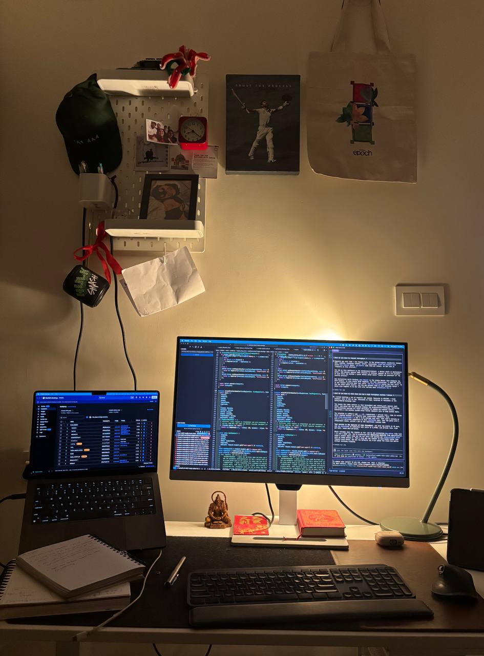 Warmly lit workspace with a monitor, laptop, desk lamp, keyboard, notebook, and a pegboard wall of personal objects